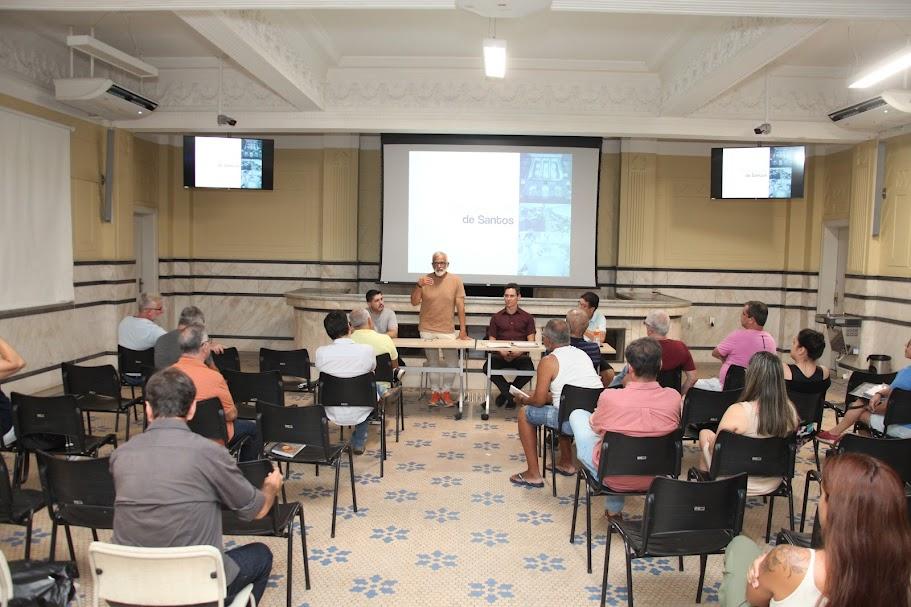 pessoas sentadas, de costas para a foto, assistindo a uma palestra. Ao fundo, uma mesa com três pessoas, sendo uma delas em pé. #paratodosverem 