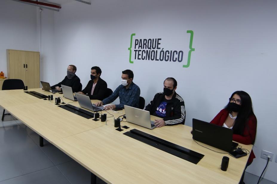 pessoas sentadas à mesa, com seus respectivos notebooks. Na parede está fixada a placa do parque tecnológico. #paratodosverem