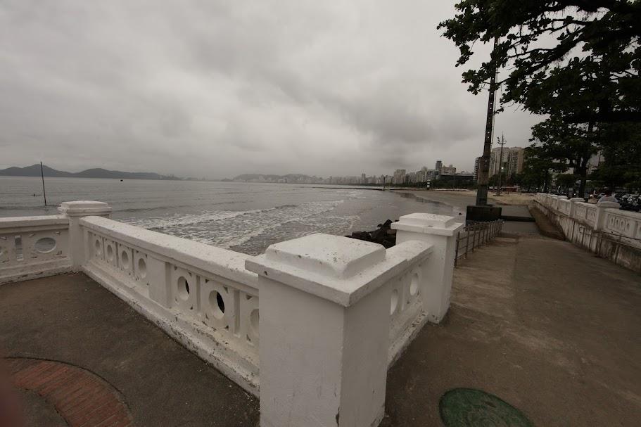 vista da faixa marítima a partir de mureta em primeiro plano na ponta da praia. O dia está nublado e o mar está calmo, com maré baixa. #paratodosverem 