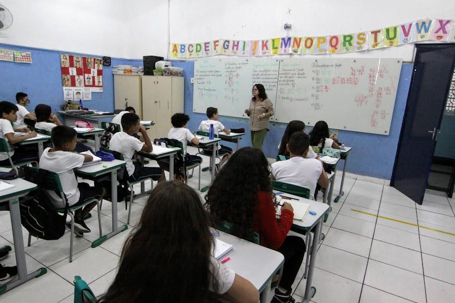 alunos estão em sala assistindo aula. Todos de costas para a foto e a professora em pé, ao fundo, de frente. #paratodosverem