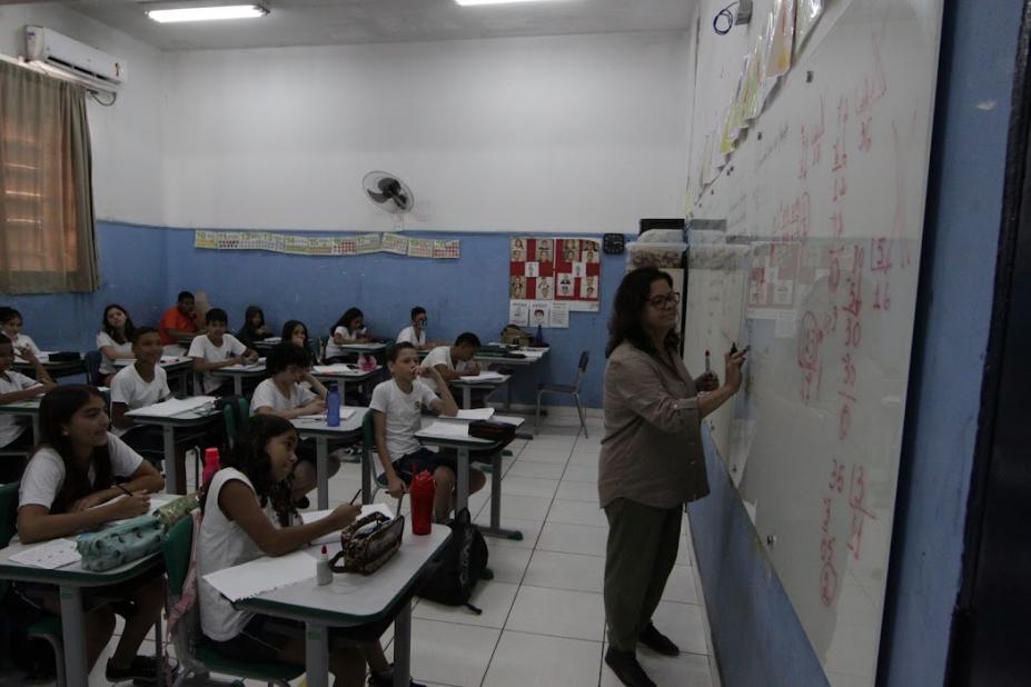 professora escreve no quadro e estudantes olham em sala de aula #paratodosverem