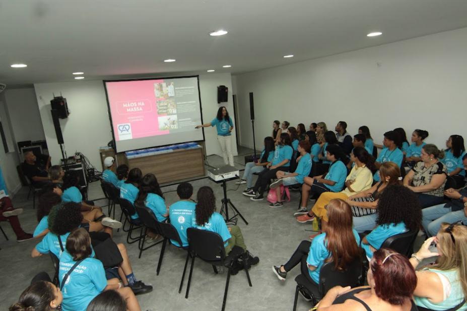 auditório com pessoas sentadas e uma delas em pé e falando à frente de um telão. #paratodosverem 