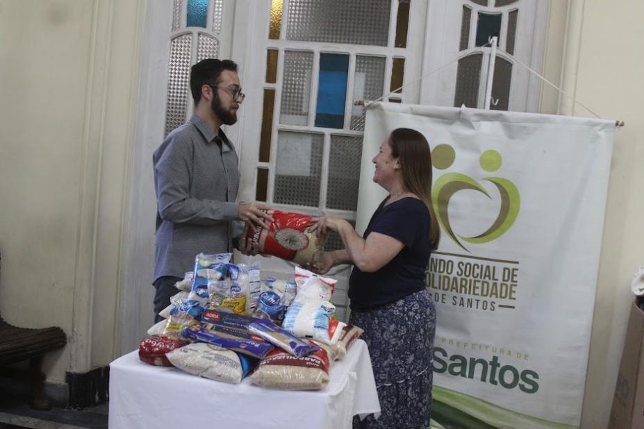 homem segura pacote de arroz ao mesmo tempo que mulher. ambos estão atrás de uma mesa com mantimentos e ao lado do banner do fundo social de solidariedade. #paratodoverem 