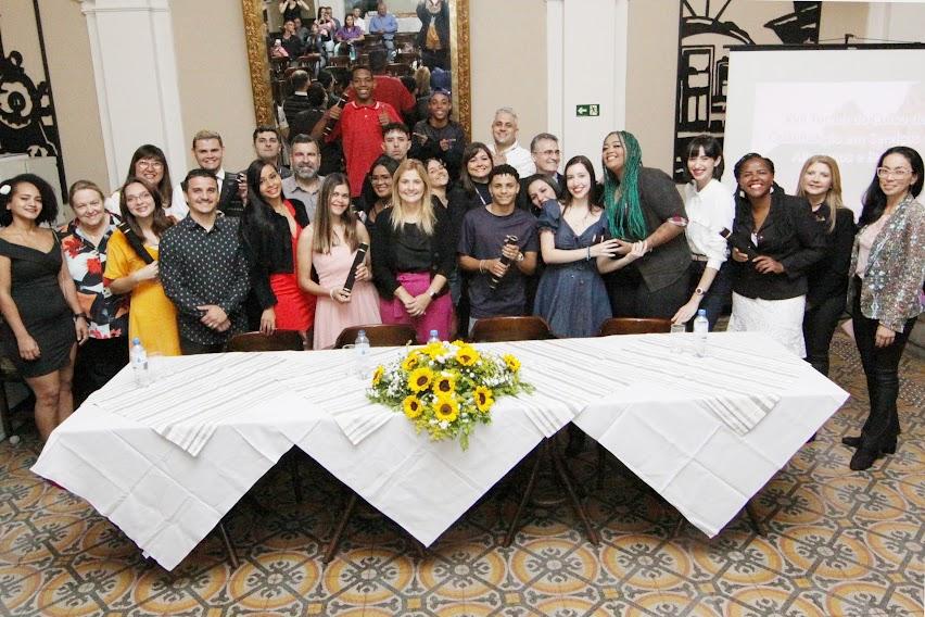 turma de alunos com dirigentes posa para foto atrás de uma mesa decorada com um buquê de flores. #paratodosverem