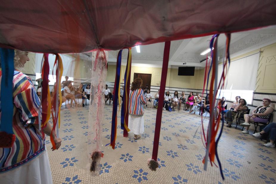 Tenda com fios coloridos em primeiro plano e mulheres sentadas em roda ao fundo. #paratodosverem 