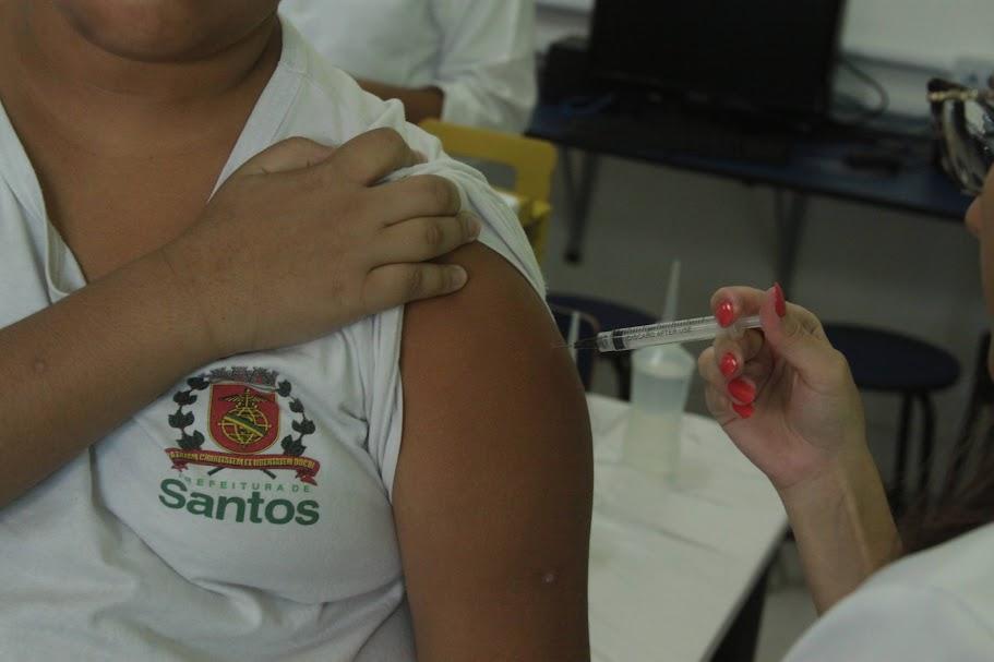 mão de mulher vacina braço de criança que usa uniforme da rede municipal. brasão de santos está no peito, à direita. criança segura a manga da camisa. #paratodosverem 
