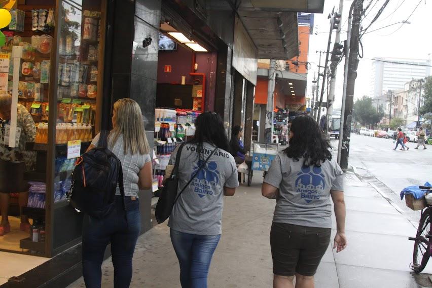 três mulheres com camiseta onde se lê nas costas amigos do bairro caminham por calçada. #paratodosverem