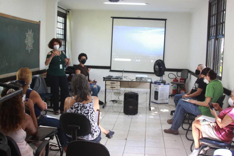 pessoas sentadas em uma sala olhando uma mulher em pé , ao fundo uma tela #paratodosverem