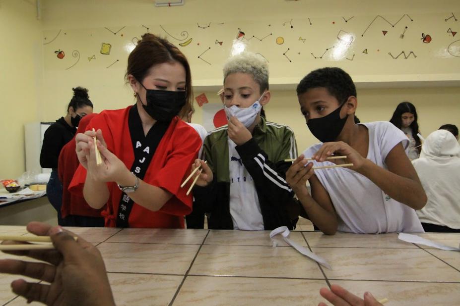 professora com vestuário japonês ensina dois alunos a segurarem hashi. #paratodosverem