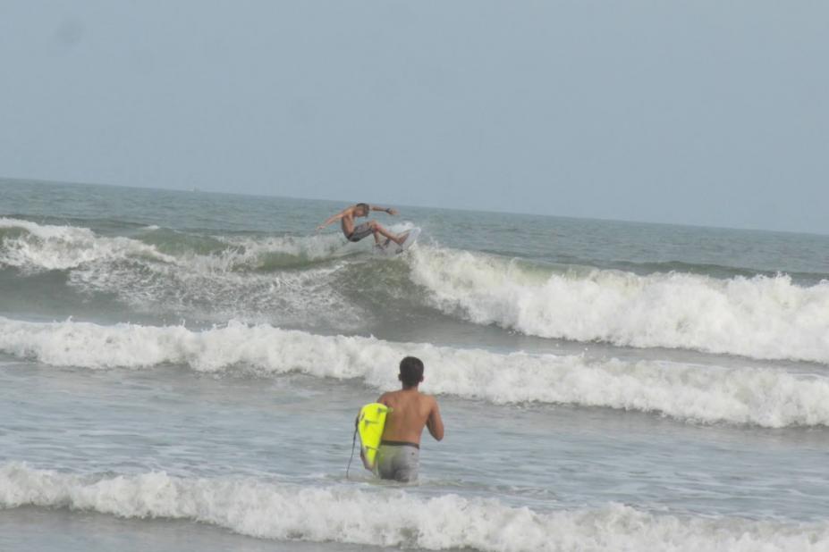 criança está entrando no mar com prancha em baixo do braço esquerdo (em primeiro plano) e outra criança surfando ao fundo. #paratodosverem 