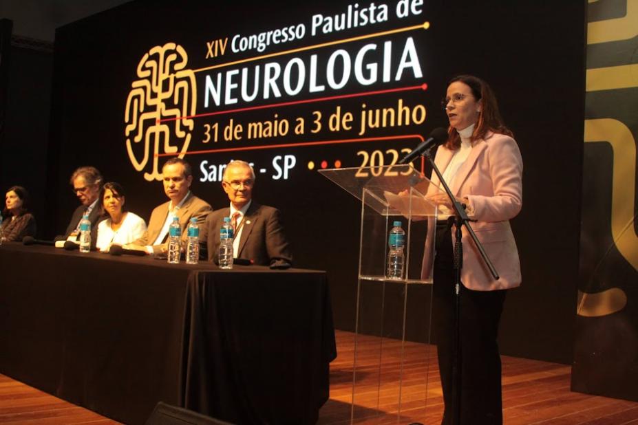 palco de evento com a vice prefeita em pé falando. cinco pessoas estão sentadas a uma mesa. #paratodosverem 