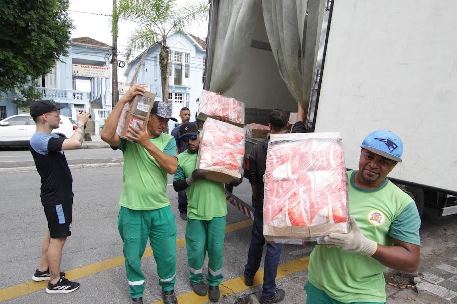 homens carregam caixas com pacotes de frango. há um caminhão atrás com a porta aberta, de onde eles retiram as caixas. #paratodosverem 