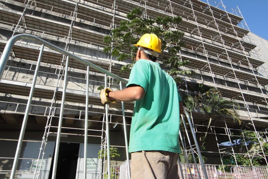 homem segura estrutura de ferro diante de fachada lateral de prédio. Ele usa capacete e luva. Está de costas para a foto. #paratodosverem 