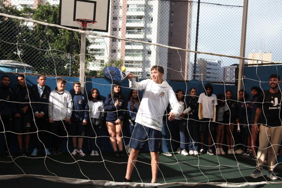 aluno joga beach tennis. ele segura raquete atrás de rede. #paratodosverem