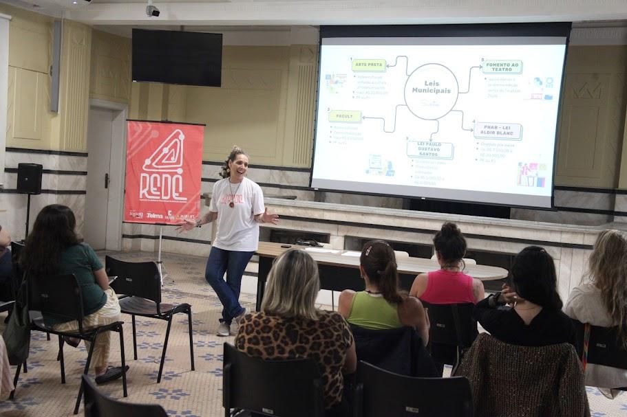 auditório com mulheres sentadas assistindo a uma palestra de uma mulher que está em pé. Há um telão ao fundo. #paratodosverem 