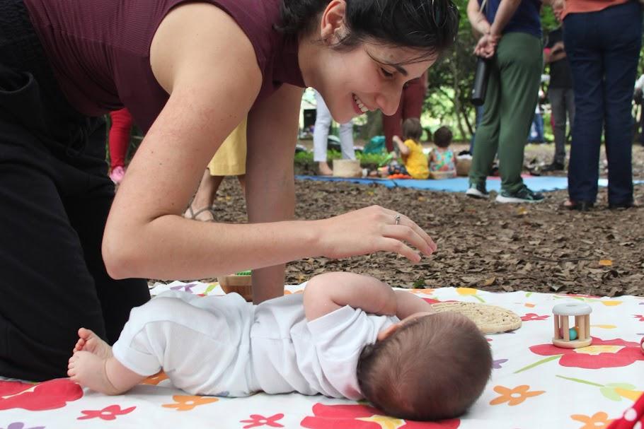 bebê está deitado no chão sobre colchonete. Mãe está ajoelhada olhando para ele e dirigindo uma das mãos ao rosto da criança. #paratodosverem 