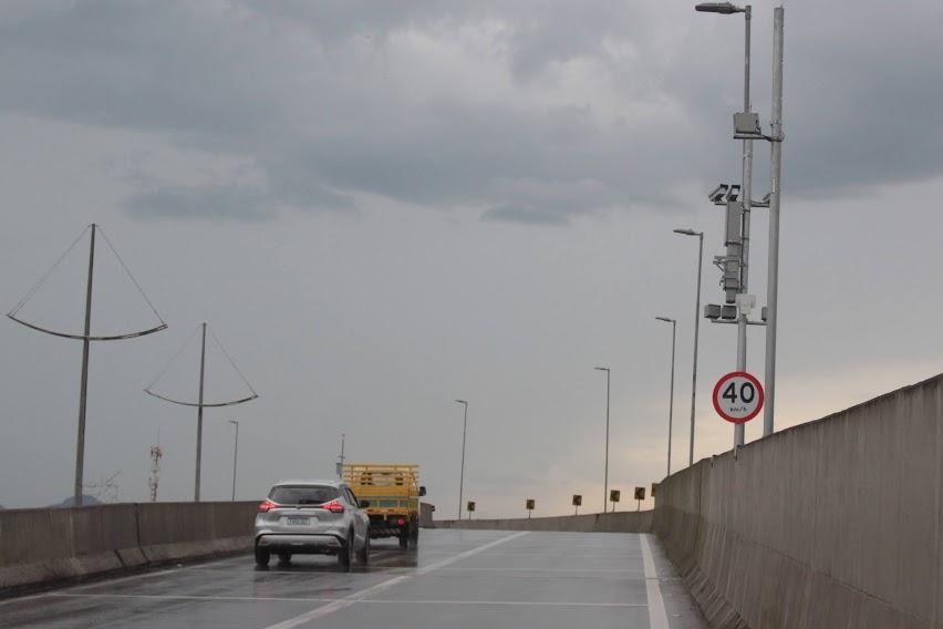 veículos trafegam em viaduto. Ao lado direito. Uma placa indicando o limite de velocidade e o radar instalado num poste. #paratodosverem