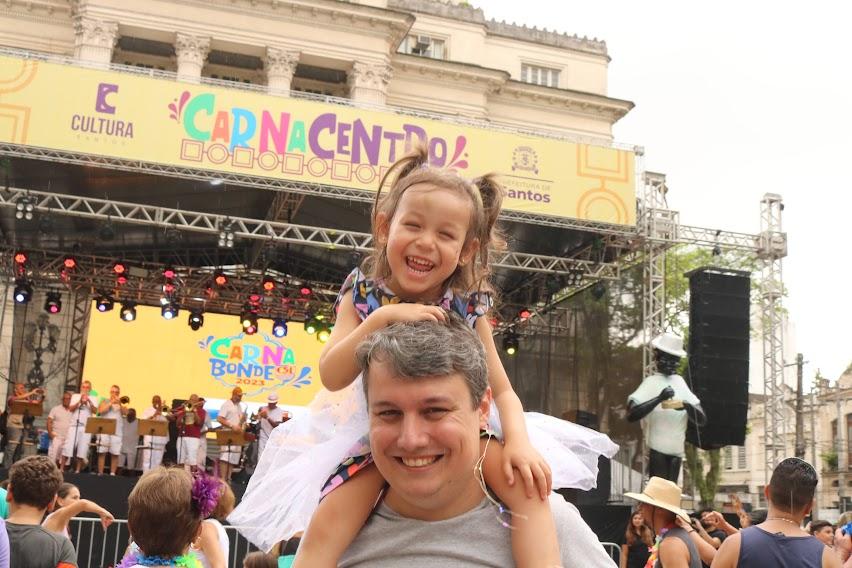 Pai com a filha no ombro, com palco do evento ao fundo. #pratodosverem