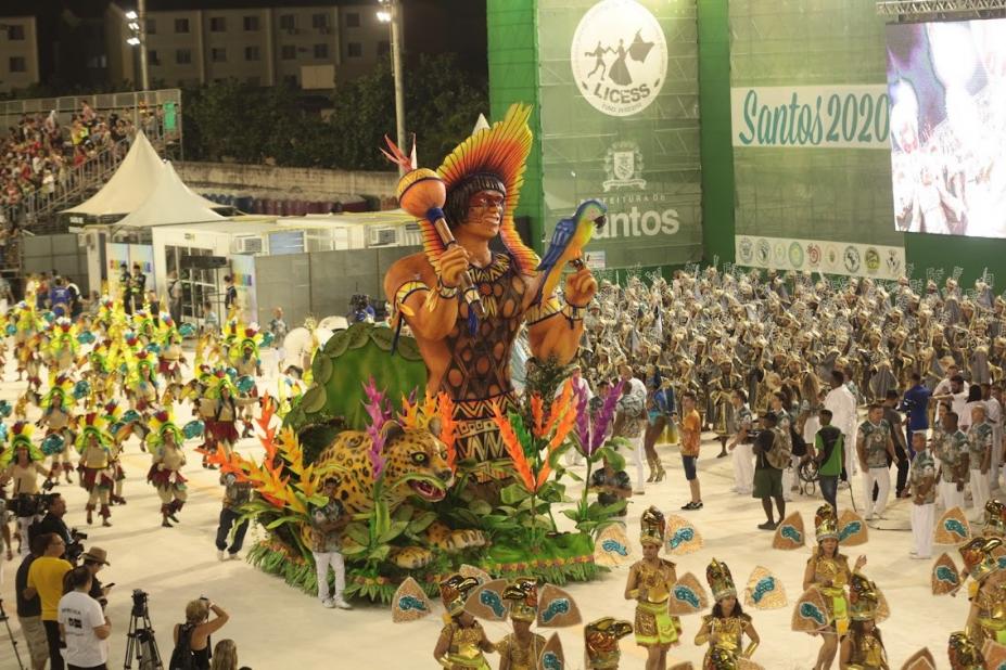 carro alegórico de indios e várias pessoas participando de desfile ao redor em passarela #paratodosverem