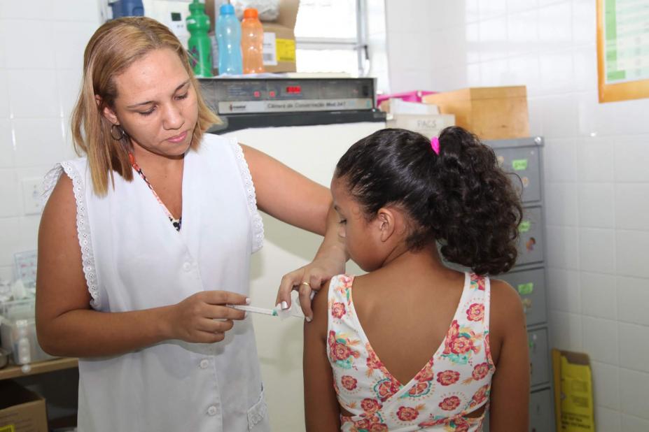 Mulher usando avental branco vacina menina que está de costas para a imagem. #Pracegover