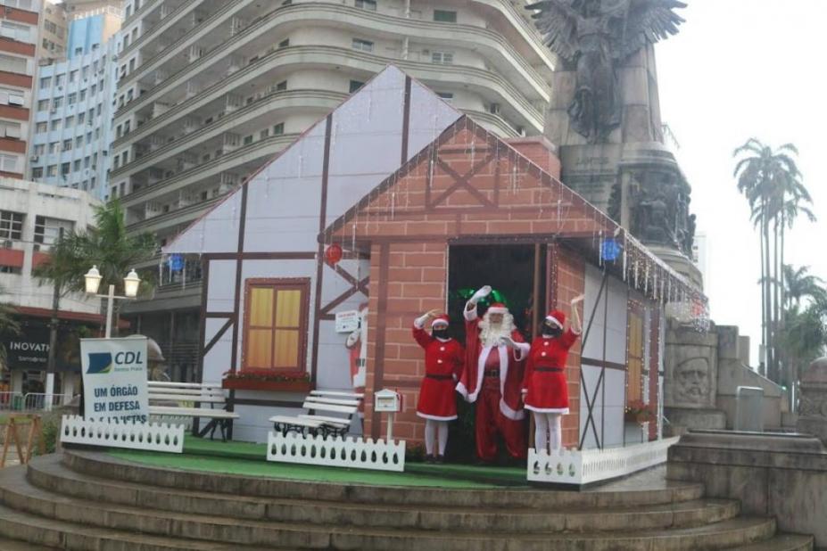 papai noel e mamães noel na frente da casinha na praça #paratodosverem