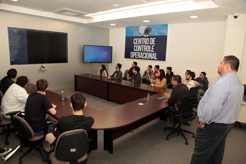 estudantes estão sentados em torno de uma mesa e diante de um telão. #paratodosverem