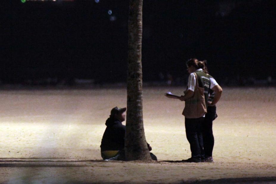 Agentes abordam pessoa em situação de rua na faixa de areia da praia, próximo a uma palmeira. A ação é realizada à noite. #Paratodosverem