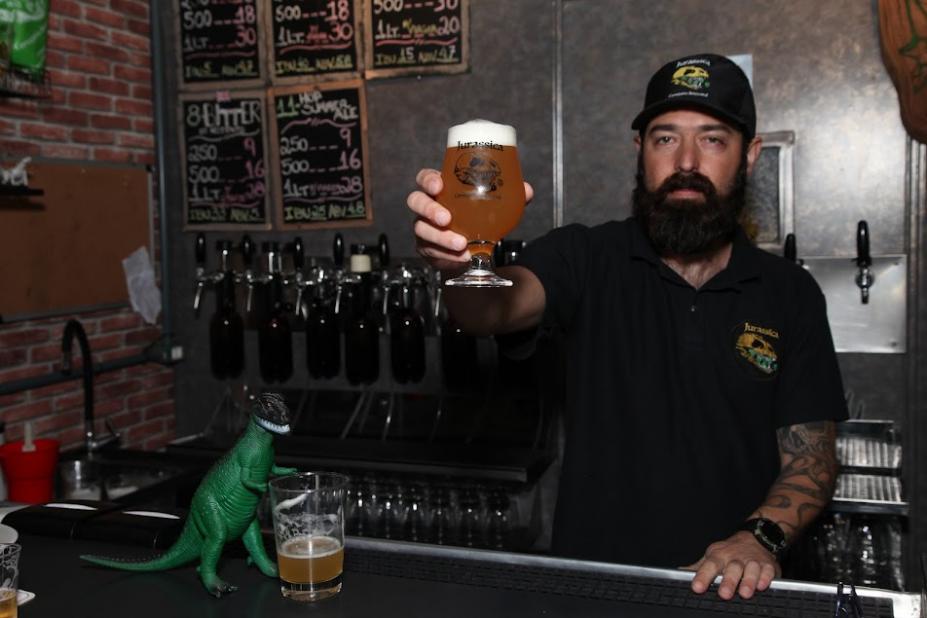 homem estende a mão e o braço com copo de cerveja cheio, como se estivesse oferecendo para a foto. #paratodosverem