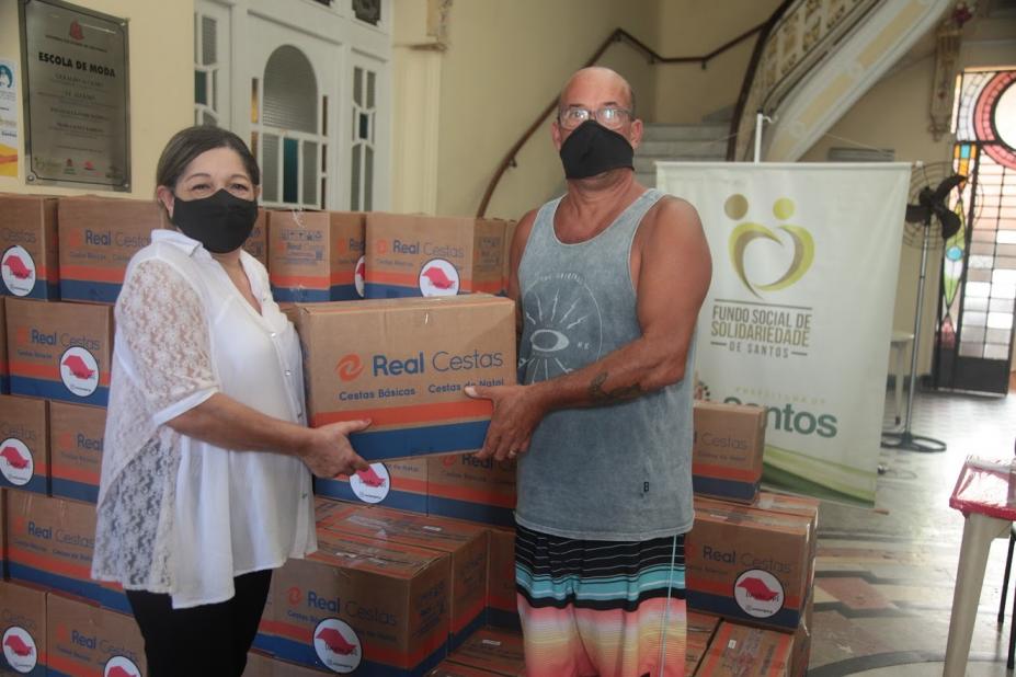 Mulher e homem estão posando para foto segurando uma caixa. Ao fundo, há diversas caixas empilhadas. #paratodosverem