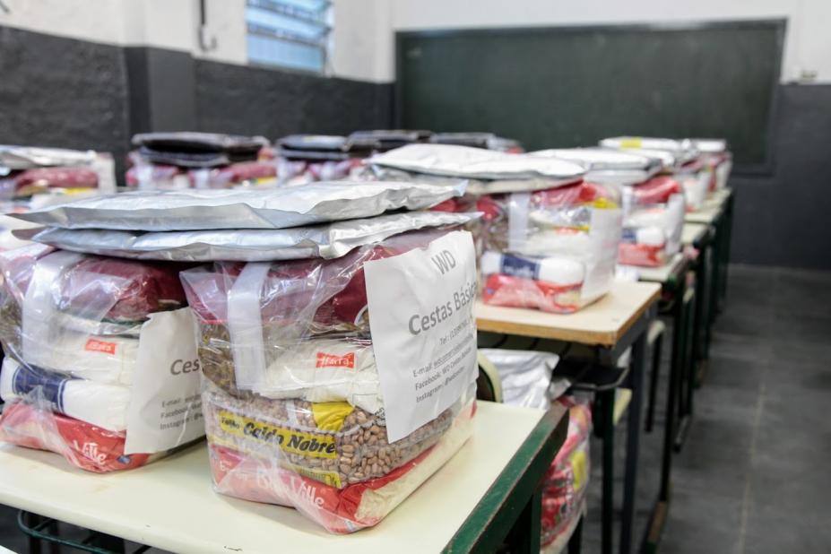 pacotes de cestas básicas estão sobre mesas escolares. #paratodosverem