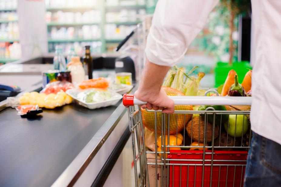 carrinho com alimentos junto à caixa de supermercado. Mãos masculinas seguram no veículo. #paratodosverem 