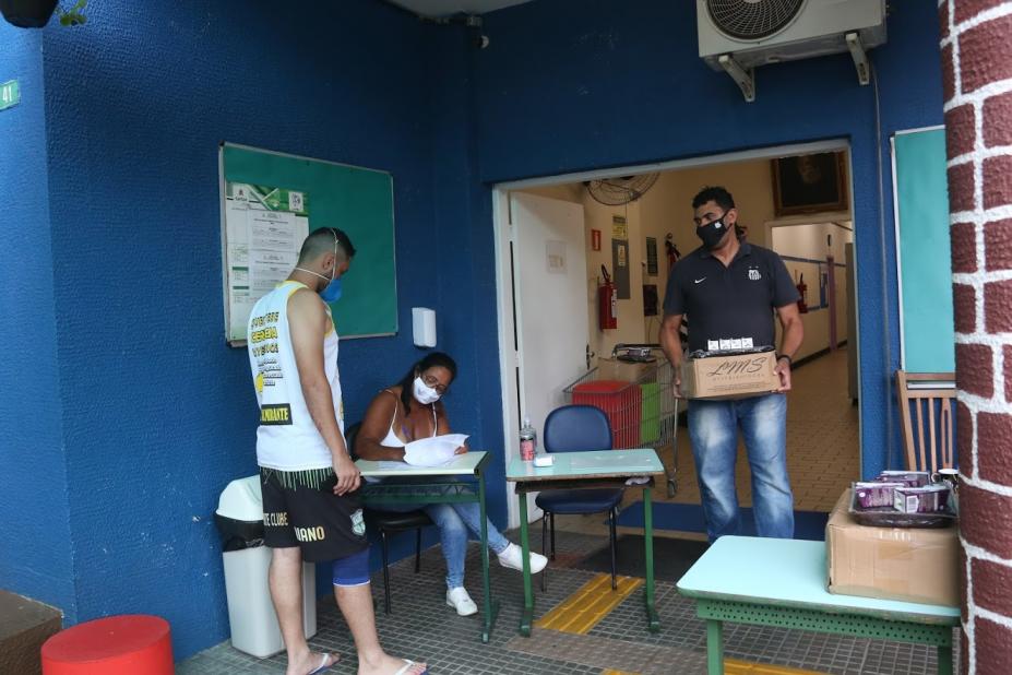 Homem está carregando um caixa, na passagem entre uma sala para outra em escola. Outro homem, à esquerda, está sendo atendido por uma mulher que mexe em papeis. #Paratodosverem