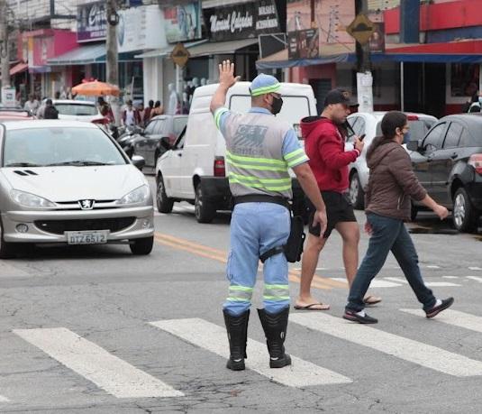 agente de trânsito está parado sobre faixa sinalizando para veículos com a mão esquerda para o alto. Ele está de costas para a foto. Dois pedestres atravessam. #paratodosverem