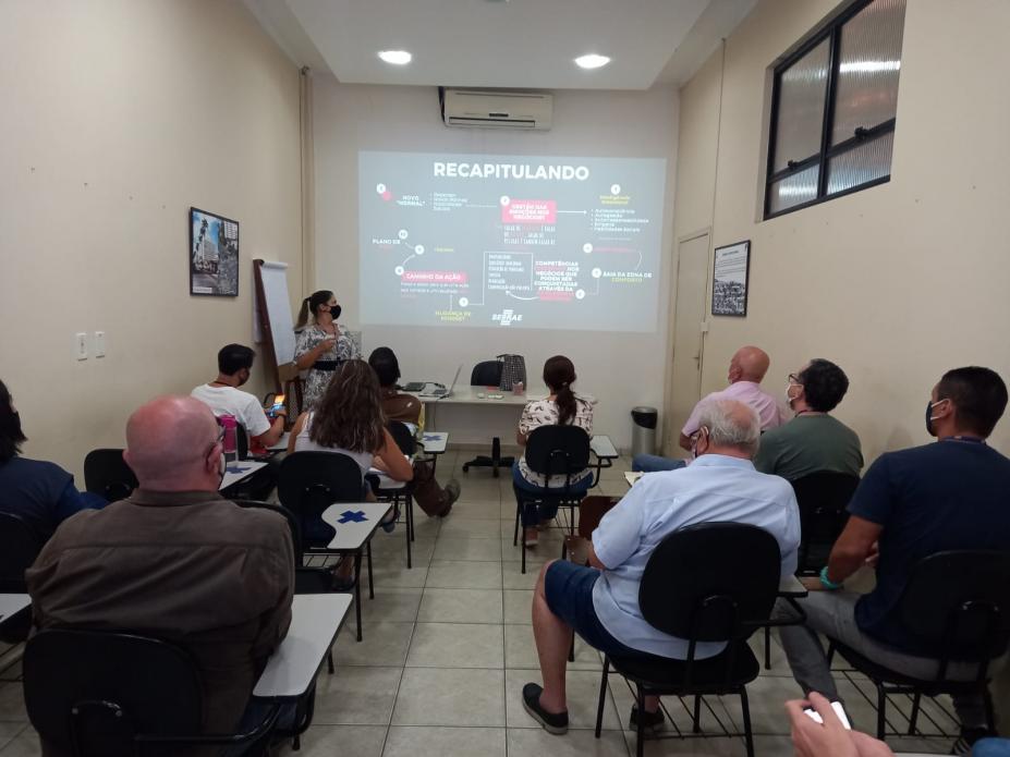 Palestrante ao lado do telão e pessoas sentadas assistindo #paratodosverem