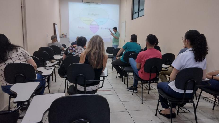 pessoas etão sentadas em cadeiras escolares, de costas para foto, assistindo palestra. #paratodosverem 