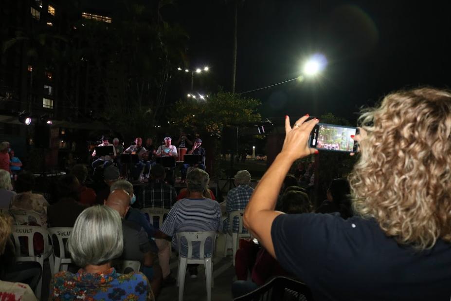 noite de apresentação musical, com músicos ao fundo. Há pessoas sentadas na plateia. Em primeiro plano, à direita, uma mulher de costas para a foto, aponta o celular para fotografar a banda. #paratodosverem