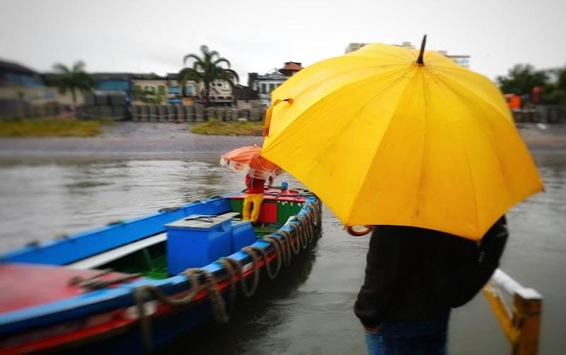 pessoa está na estação de catraias parada do lado direito segurando um guarda-chuva. Ela está de costas. Ao lado esquerdo está chegando uma catraia com uma pessoa dentro. O dia está nublado. #paratodosverem 