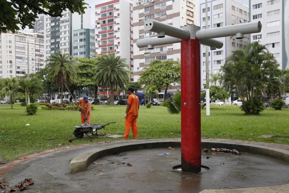 chuveirinho em primeiro plano e dois homens ao lado de carrinho de mão. Eles se movimentam para limpar caixa de deposição de areia. #paratodosverem