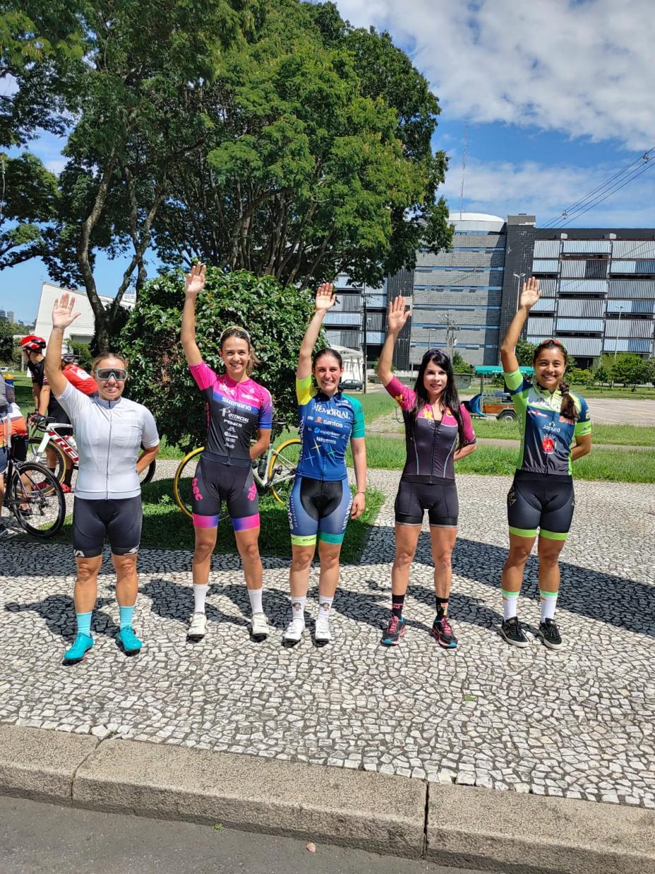 cinco mulheres estão em pé em uma calçada. Todas estão com o braço direito para o alto. Taise é a do centro. #paratodosverem