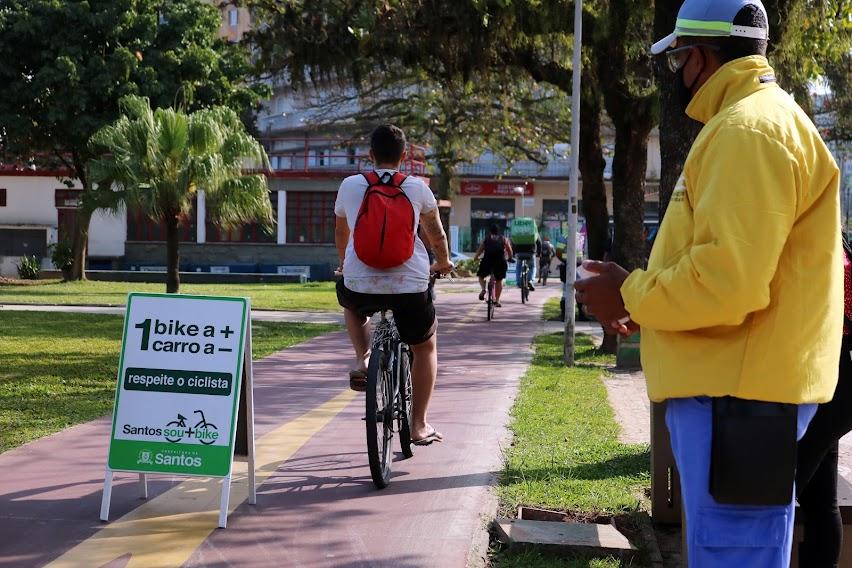 Agente de trânsito observa passagem de ciclista na ciclovia ao lado de placa com a inscrição Uma bike a mais Um carro a Menos. #pracegover