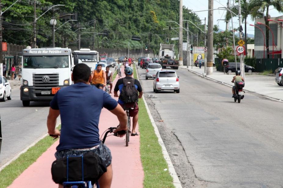 ciclistas pedalando em ciclovia. Carros estão rodando nas duas pistas entre a ciclovia. Uma pista vai, outra vem. #paratodosverem