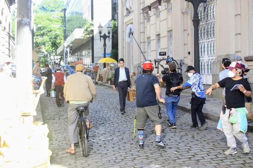 várias pessoas se movimentando em rua do centro para fazer uma filmagem. Alguns seguram equipamentos como câmeras e microfones. Outros estão com roupas antigas. #paratodosverem