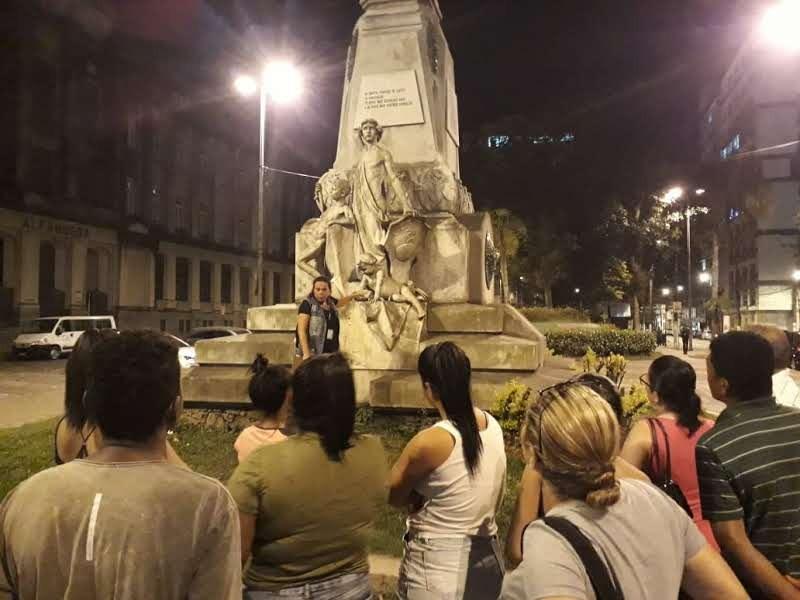 Várias pessoas estão em torno da estátua de Brás Cubas. Uma mulher orienta o grupo. #Paratodosverem