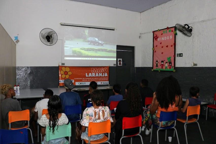 jovens estão sentados em cadeiras, de costas para a foto, assistindo a uma projeção num telão na parede. #paratodosverem