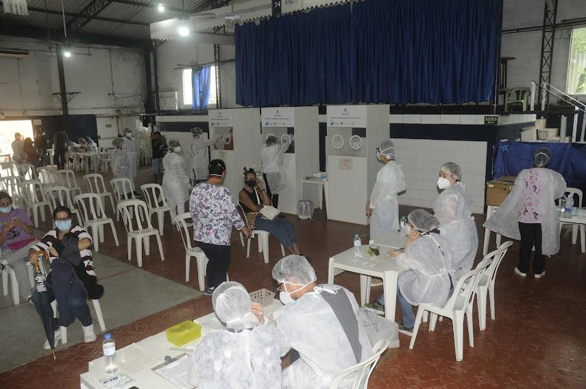 atendentes estão sentados atrás de mesas aguardando o público. Ambiente tem várias cadeiras posicionais umas distantes das outras. ao fundo, três cabines para testes PCR de covid-19. #paratodosverem