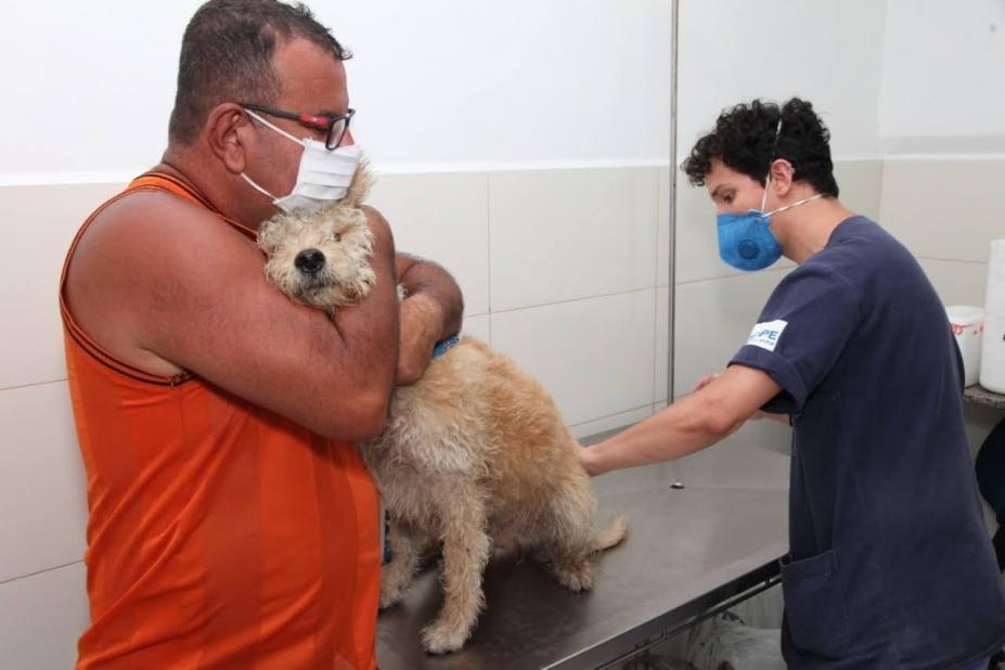 Homem está em pé abraçado a uma cachorro em atendimento veterinário. Outro homem toca nas costas do animal. #Paratodosverem