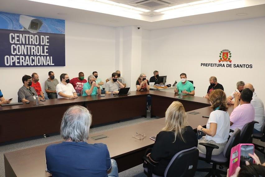 Prefeito, secretários e comerciantes sentados em mesa no formato de U, com inscrições nas paredes: Prefeitura de Santos e Centro de Controle Operacional. #pracegover