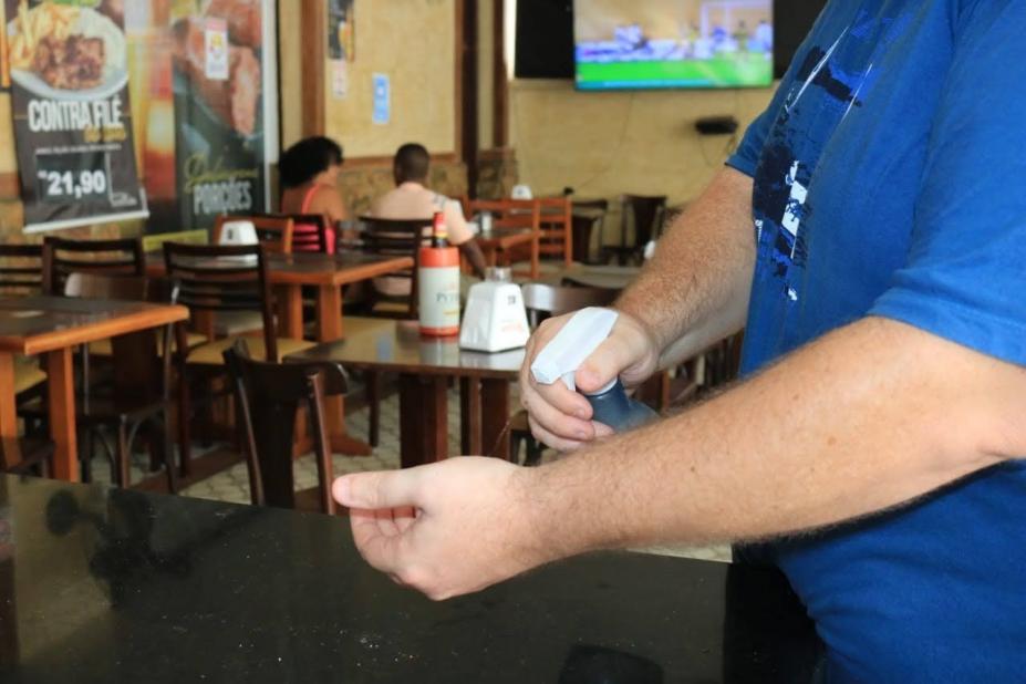 Homem borrifa álcool em uma das mãos. Ao fundo estão mesas de um restaurante. #paratodosverem