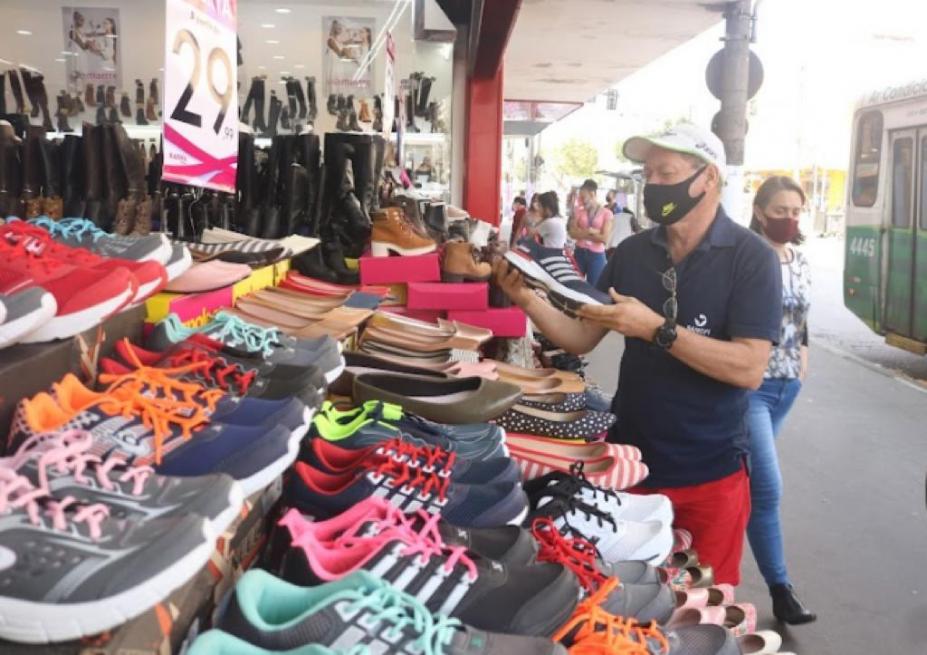 frente de loja com inúmeros pares de sapato em exposição. Um homem segura um pé de tênis. A rua está ao lado direito. #paratodosverem