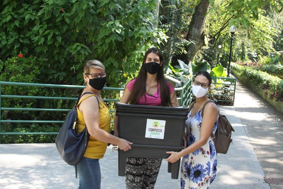 três mulheres seguram uma caixa de compostagem com vegetação ao fundo #paratodosverem